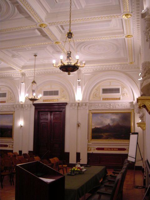 Room of the event: podium, speaker table, screen, rows of chairs, paintings on the wall, carved ceiling, etc.
