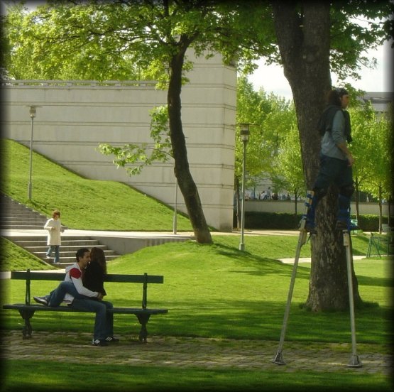 Lovers facing each other on a bench, unfazed as a man passes by them on stilts