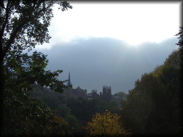 Tower of the Hub, Lawnmarket visible between trees of Princes Street Gardens, clouds and sun showing up