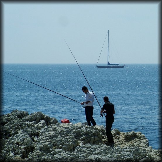 Two men fishing on the rocks, a sailing boat in the background