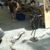 A woman stooping over some basket, a metal chicken and thin metal man