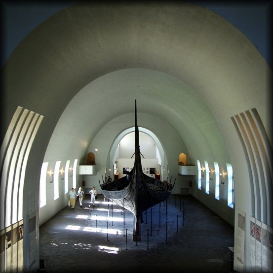 People look small as they walk by the Oseberg Ship at the Viking Ship Museum.  <em>The Oseberg Ship was found in a large burial mound at the Slagen farm in Vestfold and excavated in 1904. The ship was built in around 815-820 A.D. and had been used as a sailing vessel for many years before it was put to use as a burial ship for a prominent woman who died in 834. The ship, built of oak, was 22 meters long and 5 meters wide. The 12 strakes were secured with iron nails. The ship was designed for both rowing and sailing. With a square sail of about 90 sq. m., it could reach speeds of over 10 knots. The top strake had 15 oar holes.</em>  <em>The skeletons of two women were found in the grave. One, aged 60-70, suffered badly from arthritis and other maladies; the second was aged 25-30. It is not clear which one was the most important in life, or whether one was sacrificed to accompany the other in death. Although the high-ranking woman's identity is unknown, it has been suggested that it is the burial of Queen Asa of the Ynglinge clan, mother of Halfdan the Black and grandmother of Harald Fairhair. This theory is now generally dispelled, and it is now thought that she may have been a priestess.</em>
