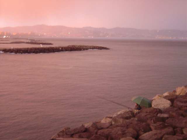 Rose light filtering through the clouds at sunset over a rose sea, fisher under green umbrella