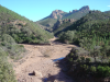 Barren reddish rocks, small pool of water, trees, mountains, clear blue sky