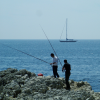 Two men fishing on the rocks, a sailing boat in the background