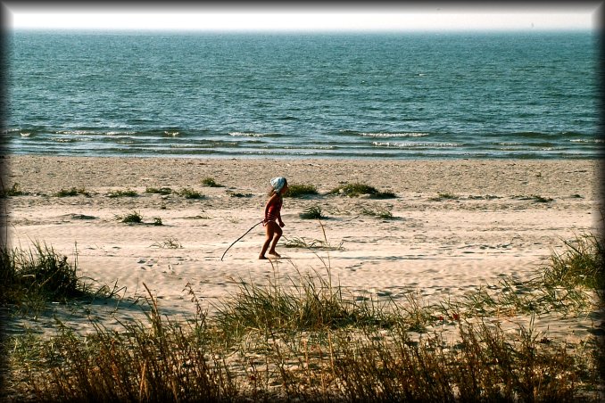 A little girl playing on her own on the beach, dragging a stick in the sand.